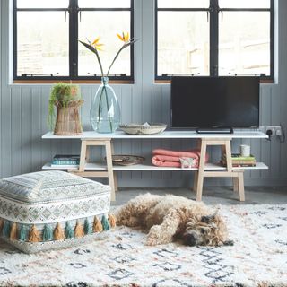 Shaggy dog laying on a fluffy rug in a living room next to an Aztec pouffe