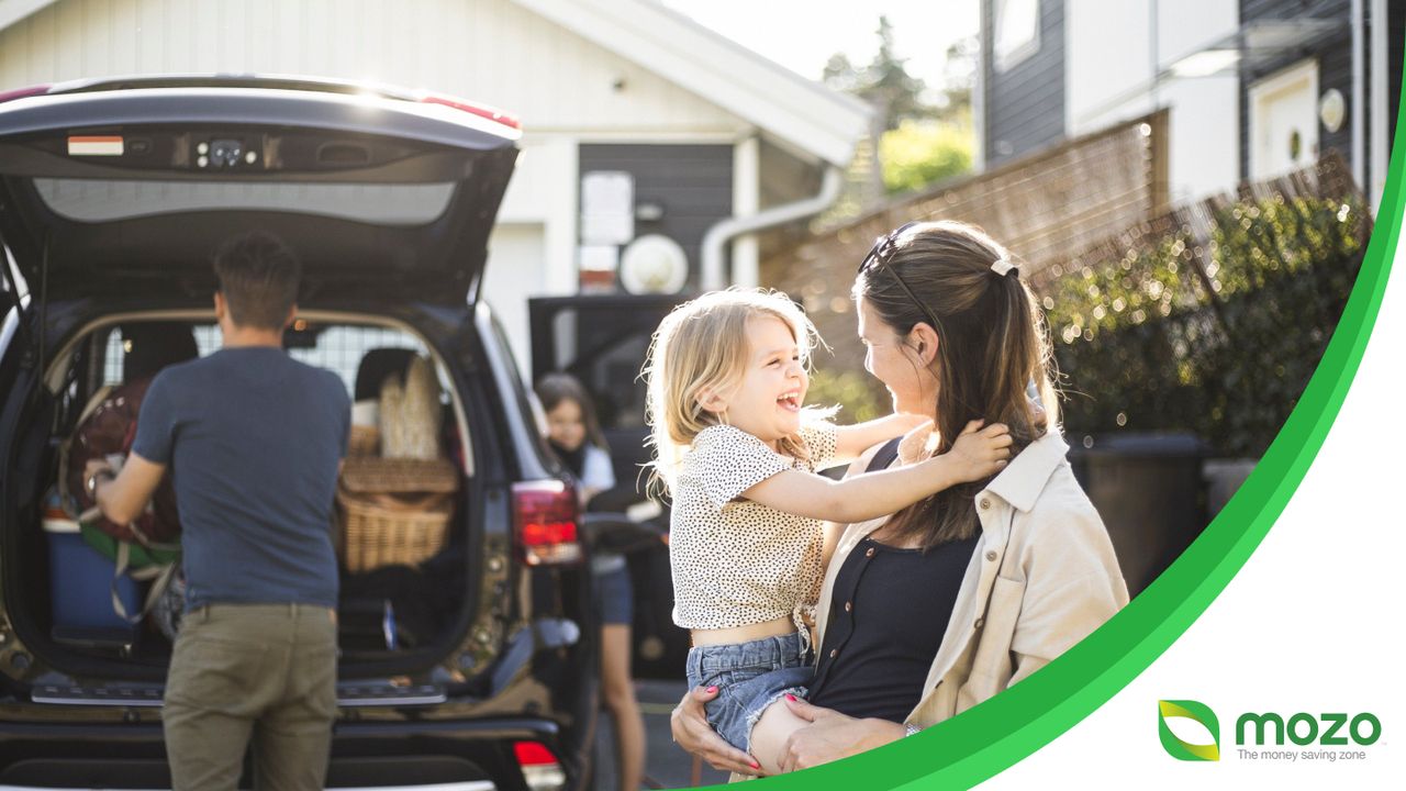 A mother holding a young daughter while a man packs the boot of a car