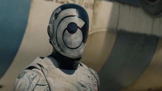 Screen grab from the trailer for Murderbot on Apple TV Plus showing a robot with a mask-like face in front of a large structure that may be a spaceship