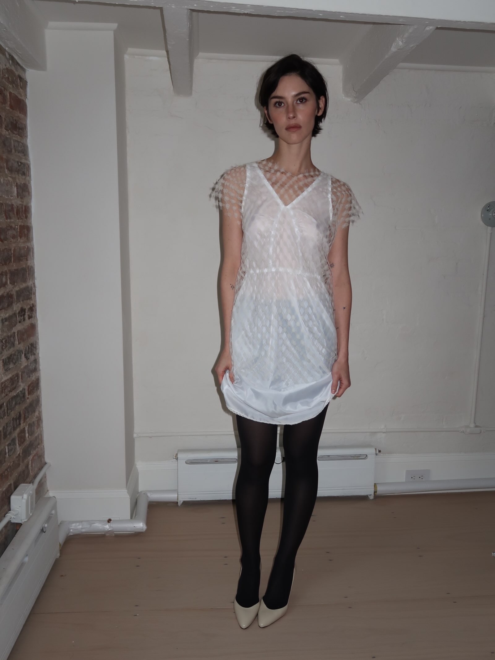 Gracie Abrams poses for a photo wearing a white semi-sheer minidress with opaque black tights and white pointed-toe heels.