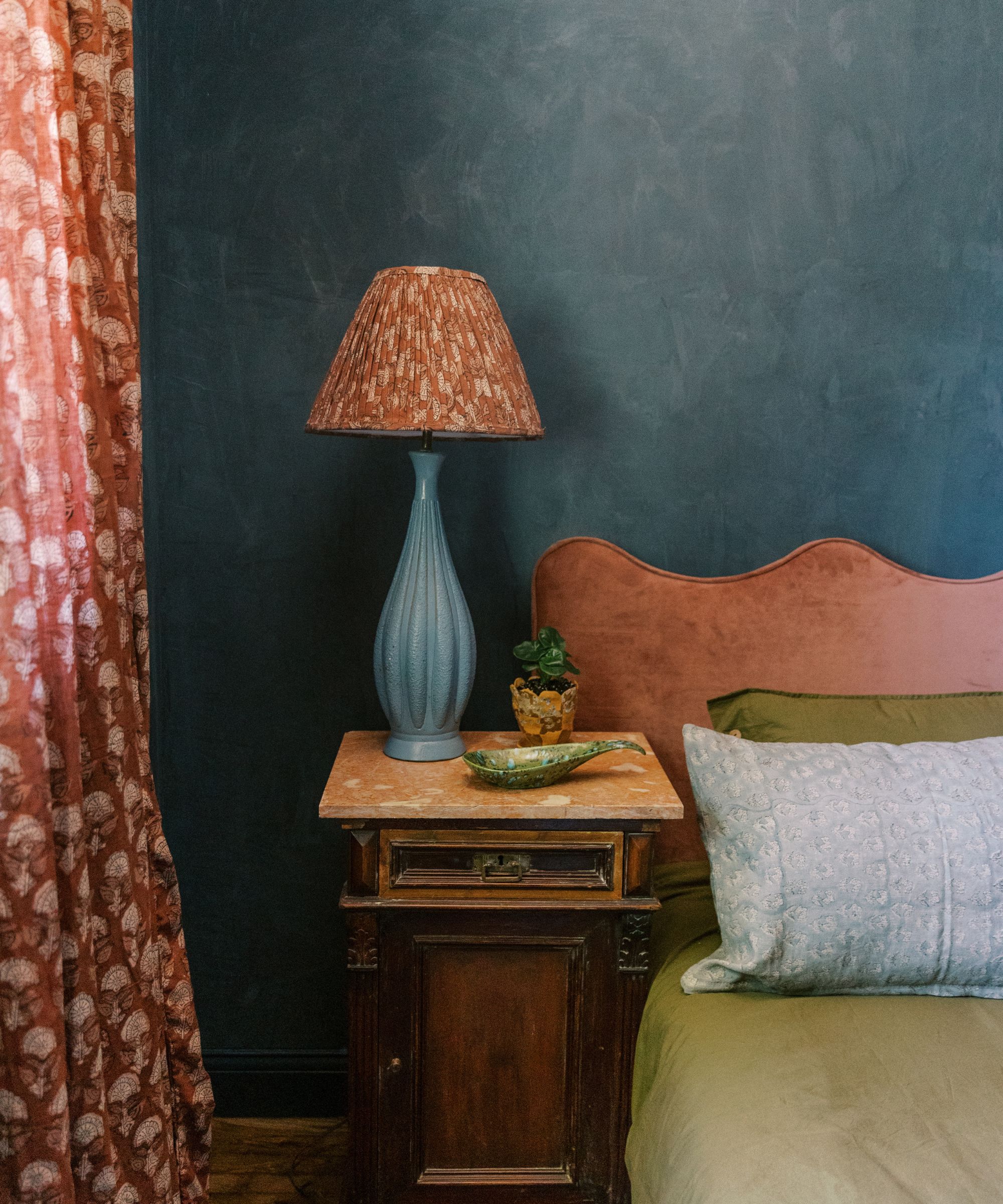 Limewashed dark blue walls in a bedroom with a pink headboard, green bedding, and a wooden nightstand with a blue lamp.