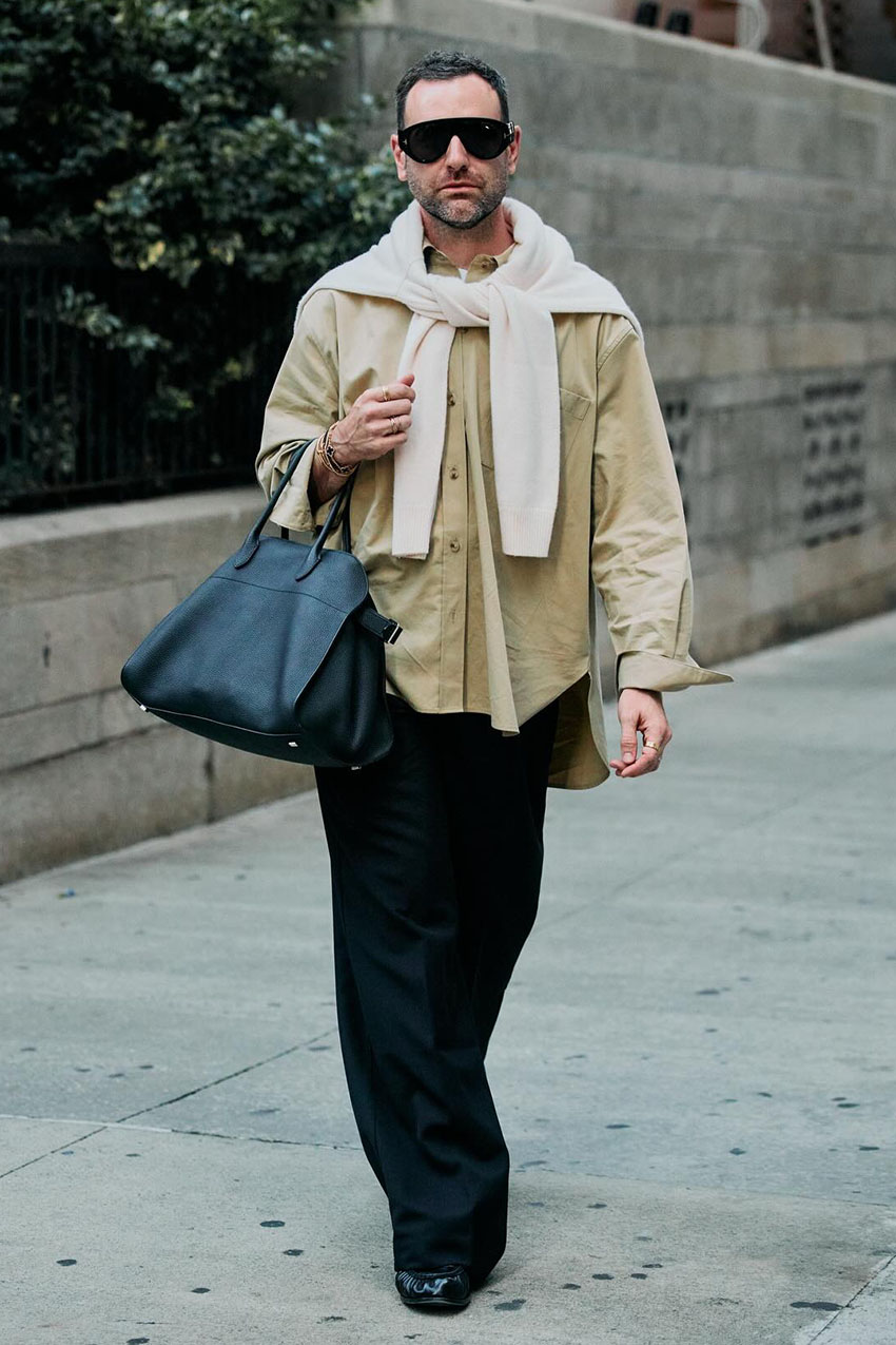 A man wearing a tan button-down, black pants, a black leather bag, and white longsleeve sweater