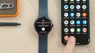 Google Pixel Watch 3 on a wood desk next to a Google Pixel 9 phone with a finger pointing to the Watch app