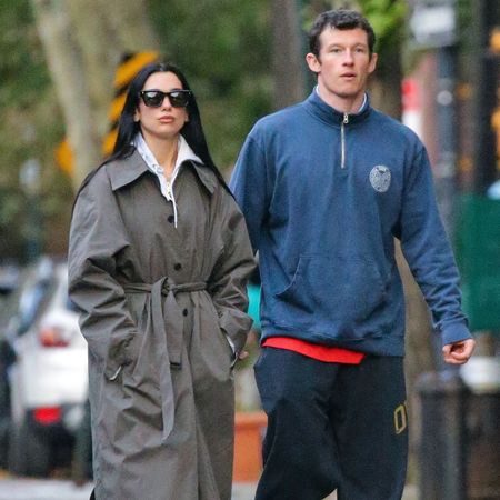 Dua Lipa walks down the street with Calum Turner wearing a grey single-breasted trench coat with leggings, a hoodie and black trainers.