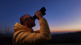 A man in a yellow jacket looking through binoculars at the sky with a sunset and crescent moon in the background.