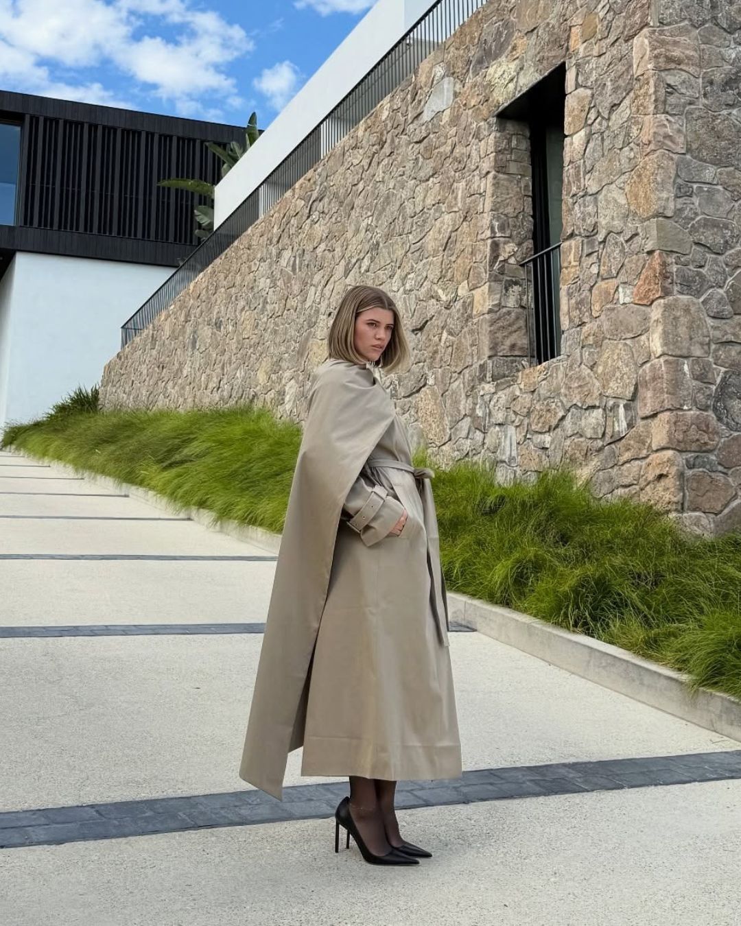 Sofia Richie poses for a photo outside wearing a long-line beige scarf coat with sheer tights and black pointed-toe heels.