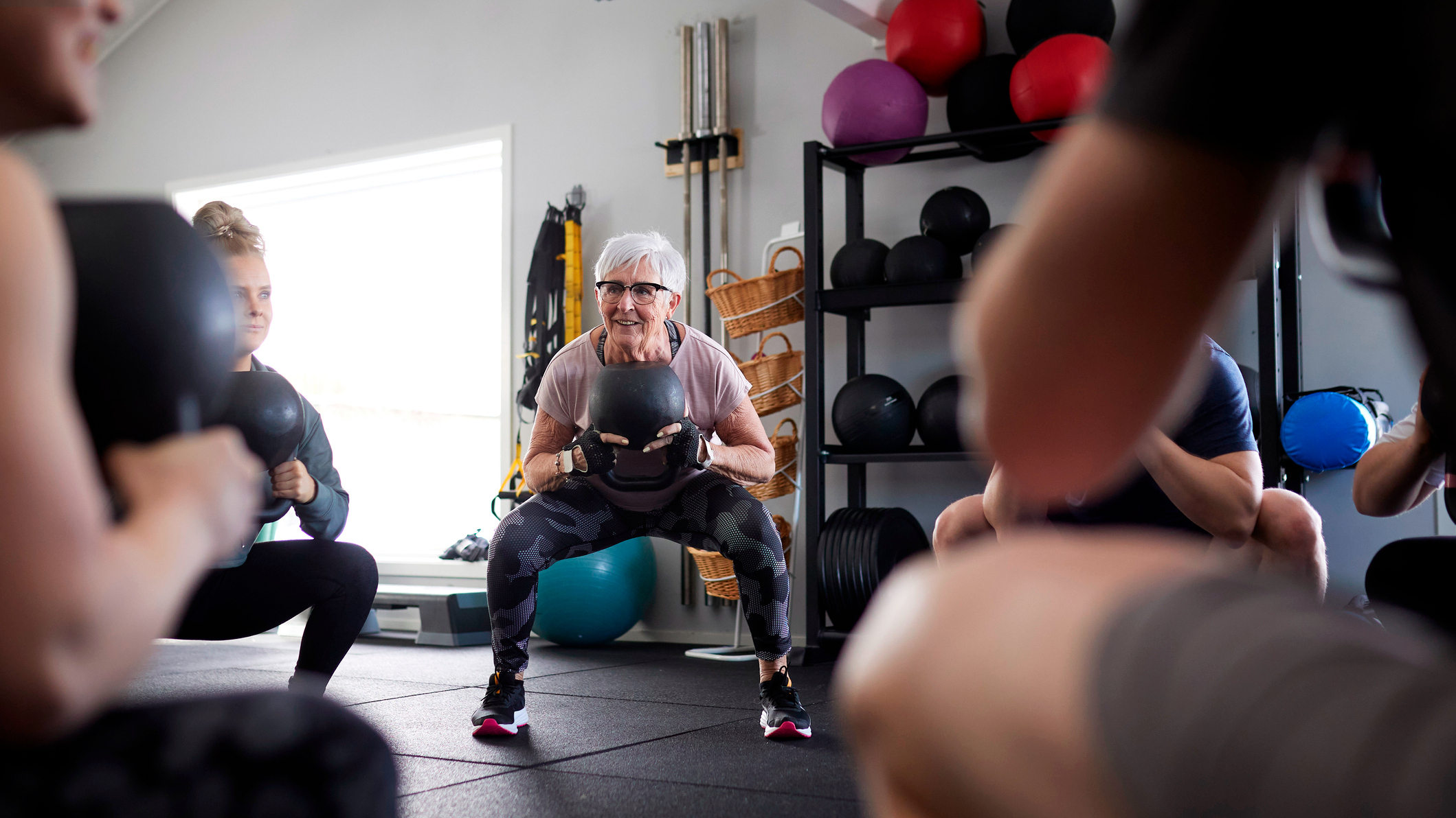 Group of people in a gym, in a circle, squatting while holding kettlebells
