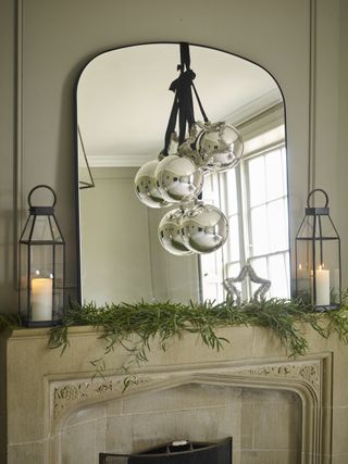 Christmas mantel with mirror, three oversized ornaments in front of mirror, lanterns each side, foliage in front on mantel