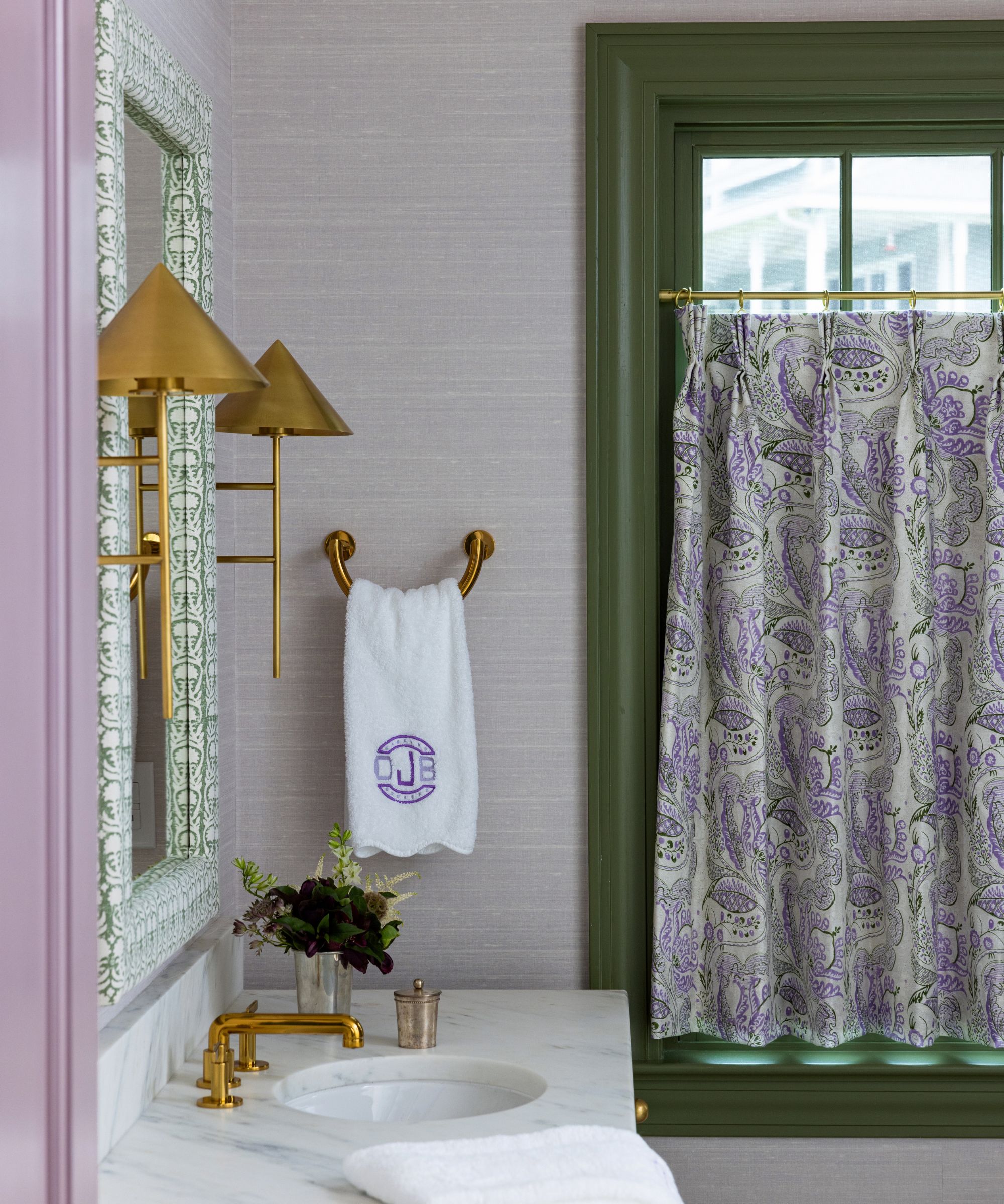 Bathroom with lilac grasscloth walls, green mirror with gold sconces, marble sink and gold tap, green painted window with purple floral curtain