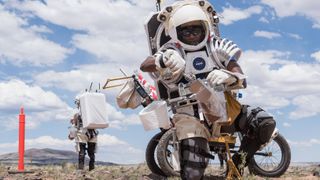 two astronauts in the field in simulated spacesuits. the one in front uses a lunar rover and tools to pick up a soil sample. clousd and blue sky are visible in the background