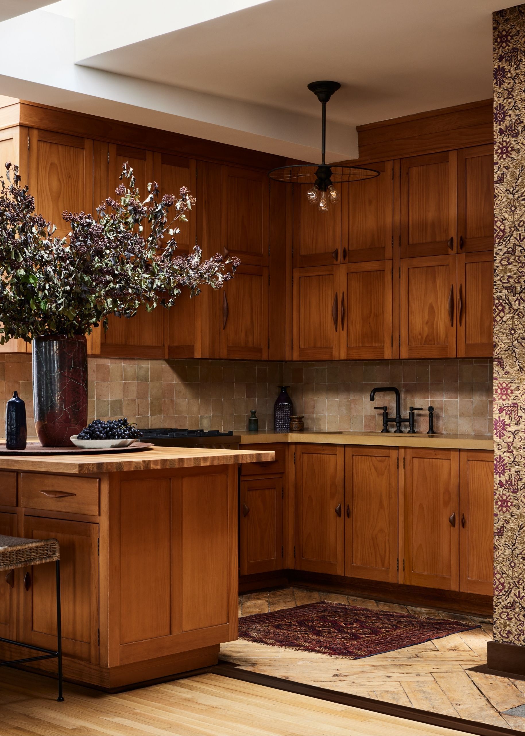 Small rustic kitchen with warm wooden cabinetry and iron hardware. There is a large black vase on the counter with dark purple flowers.