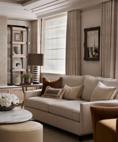 Neutral, cozy living room with cream sofa, curtains, walls and carpet, brown armchair, cushions, shelving and artwork, ottoman and coffee table in front of sofa
