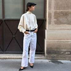 andrea wears bright white straight leg pants, a voluminous turtleneck blouse with black pointed toe shoes a black belt and a black hat