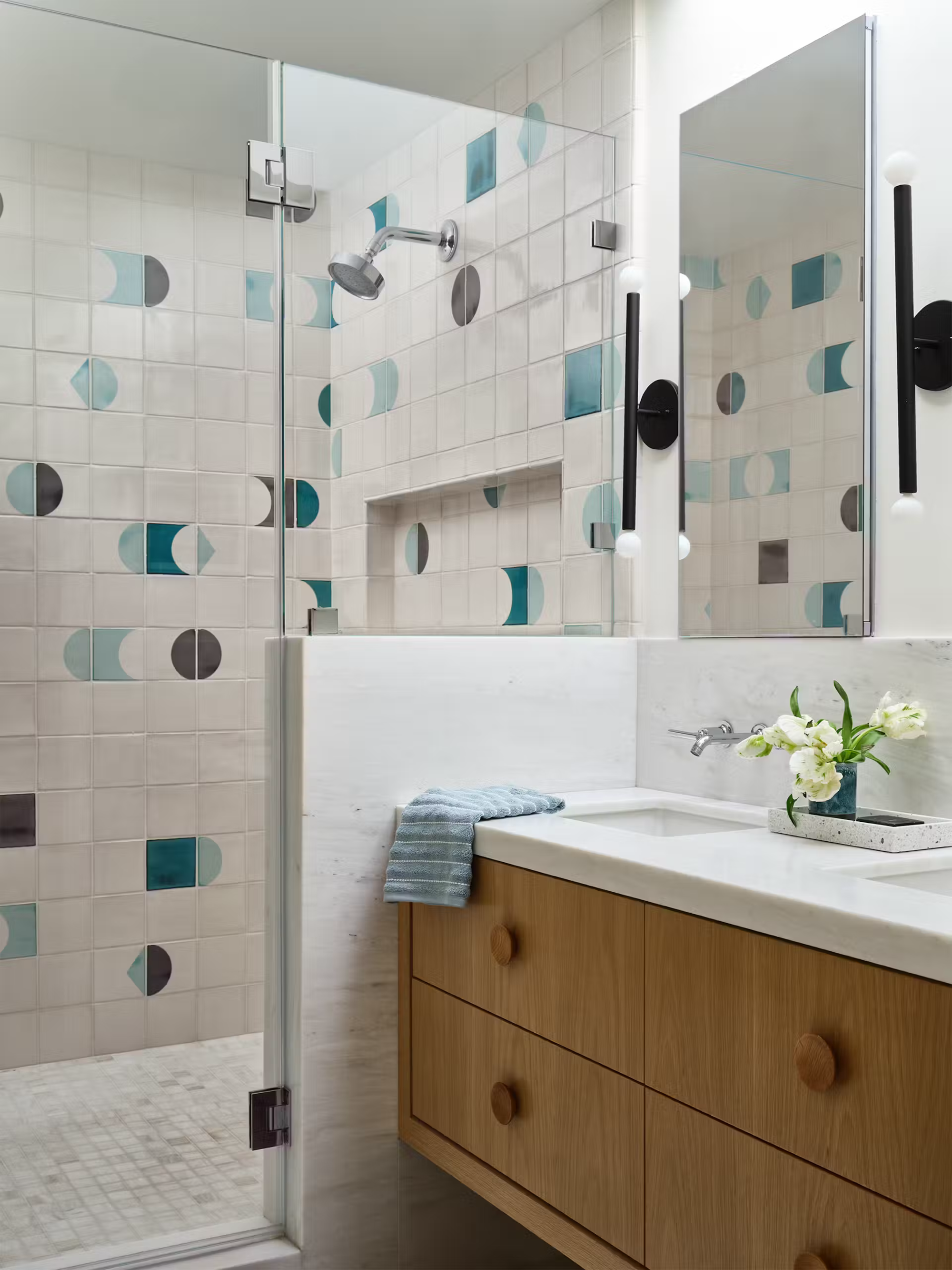 mid century modern bathroom with patterned tiles, timber vanity, marble counter, built-in sinks, mirrored cabinets, nib wall separating shower, and wall sconce