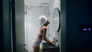 A woman with a towel on her head washing face at bathroom sink looking in mirror