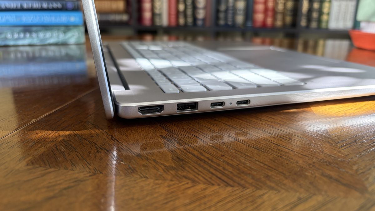 A silver OmniBook 3 laptop viewed from the side, displaying several ports, sits on a wooden table with a book-filled background.