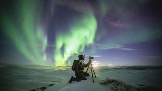 Photographer with tripod watching the green Aurora Borealis sitting in the snow, Honningsvag, Nordkapp, Troms og Finnmark, Norway