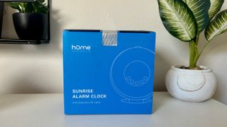 Blue box of HomeLabs sunrise alarm clock next to plant on white surface