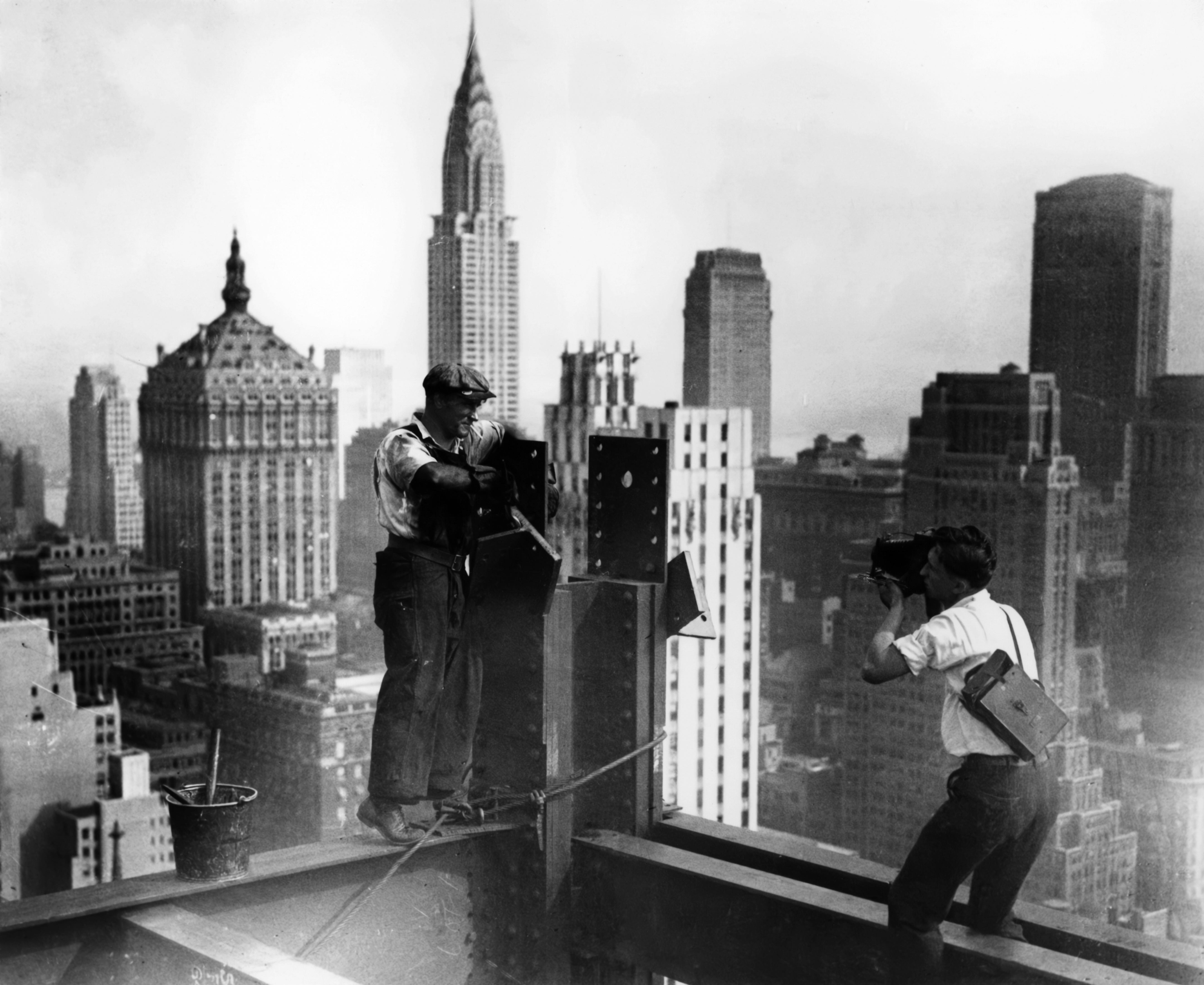 A photographer crouches on a steel beam to shoot an ironworker posing on the structure high above New York, the Chrysler Building prominent among the skyscrapers stretching into the haze below.