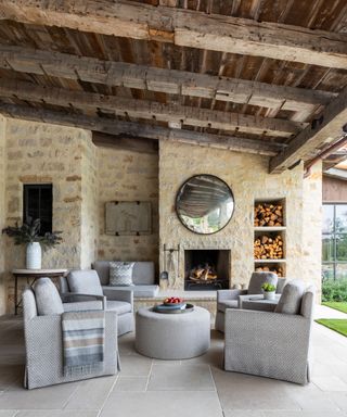Outdoor porch and verandah area, rustic reclaimed dark wood ceiling, light stone wall, outdoor furniture - four gray armchairs, rounded gray ottoman, gray stone flooring, log storage on wall, rounded mirror on wall, alcove seat