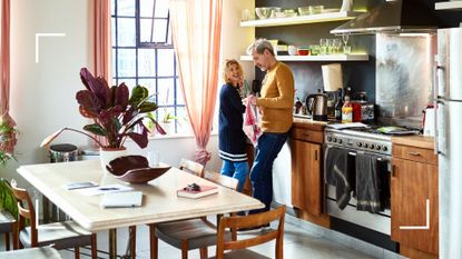 Couple talking together in the kitchen with view of dining room table with notebooks on, representing how to lower mental load in a relationship