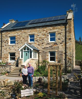 male and female couple stood outside home with a rustic stone finish and solar panels on roof