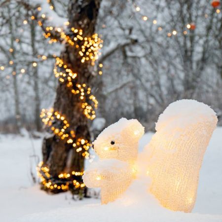 A lit up squirrel statue in the snow under a tree decorated with lights and ornaments