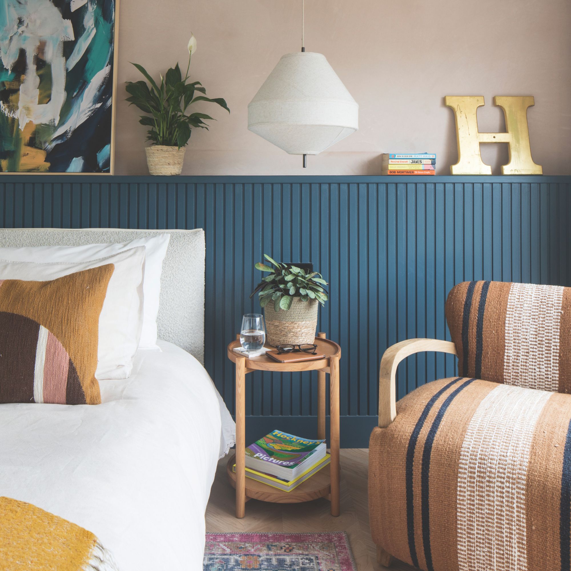 Pink painted bedroom with blue half panelling behind a white bed, and a bedside table and accent chair next to it