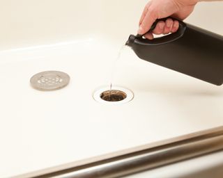 Man pouring cleaning liquid from a black bottle down a shower drain with its steel grate removed