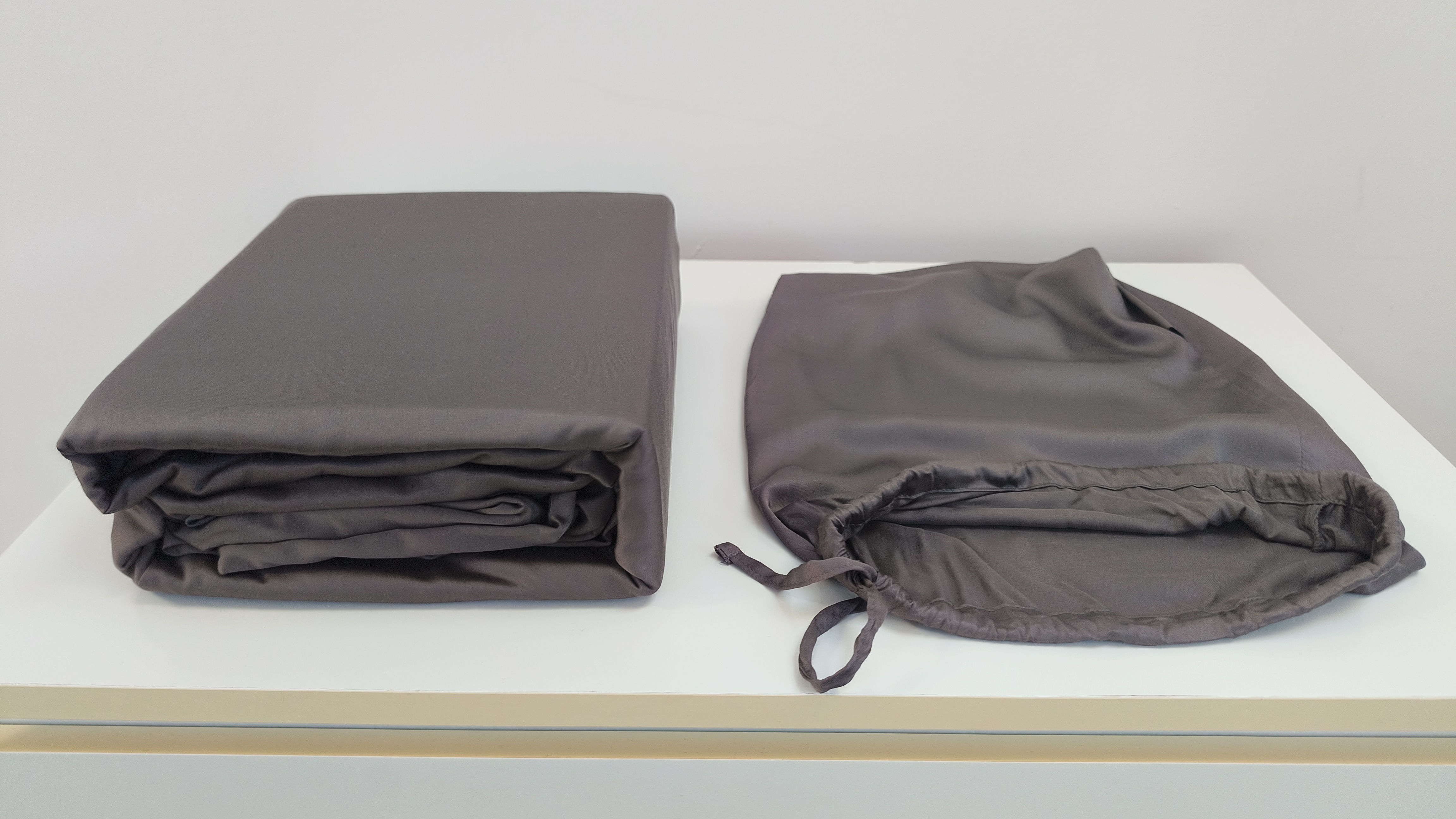A neatly folded stack of dark grey bedding sits next to its empty, matching drawstring storage bag on a clean white countertop.