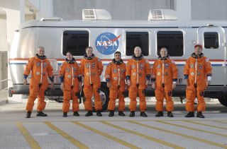 seven astronauts in orange flight suits stand in front of a silver airstream trailer.