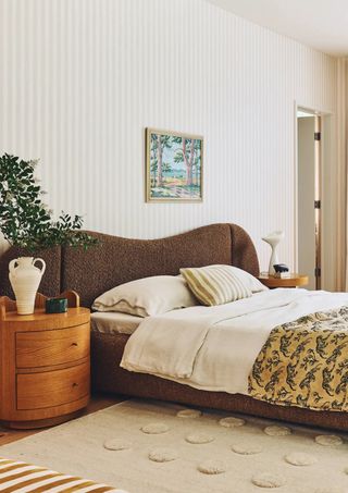 A neutral bedroom with a bed that has a brown, extended headboard. There is an orange-toned wood side table with a white vase on it beside the bed. The walls of the room are a striped wallpaper