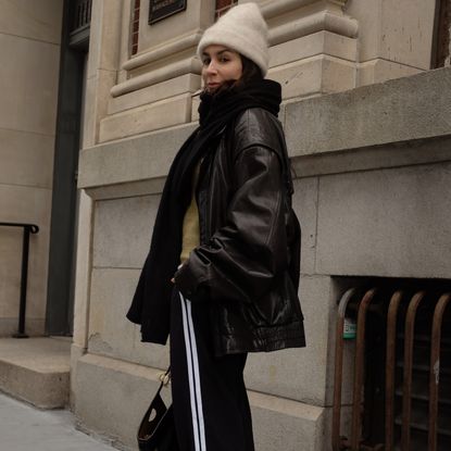 Lauren caruso looking over shoulder wearing white beanie, scarf, leather jacket, track pants 