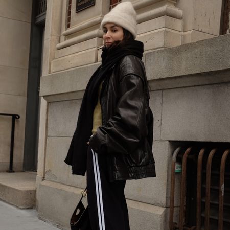 Lauren caruso looking over shoulder wearing white beanie, scarf, leather jacket, track pants 