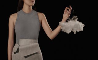View of a woman in a grey sleeveless outfit with her hand above a puff of smoke resembling a cloud in a space with a black background