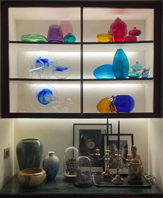 a lit display cabinet filled with colorful glass-blown vases above a shelf with our displayed items such as pictures, vases and candles