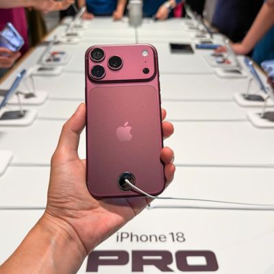 An iPhone 18 Pro smartphone is held above a branded display table as customers examine phones on either side inside an Apple Store