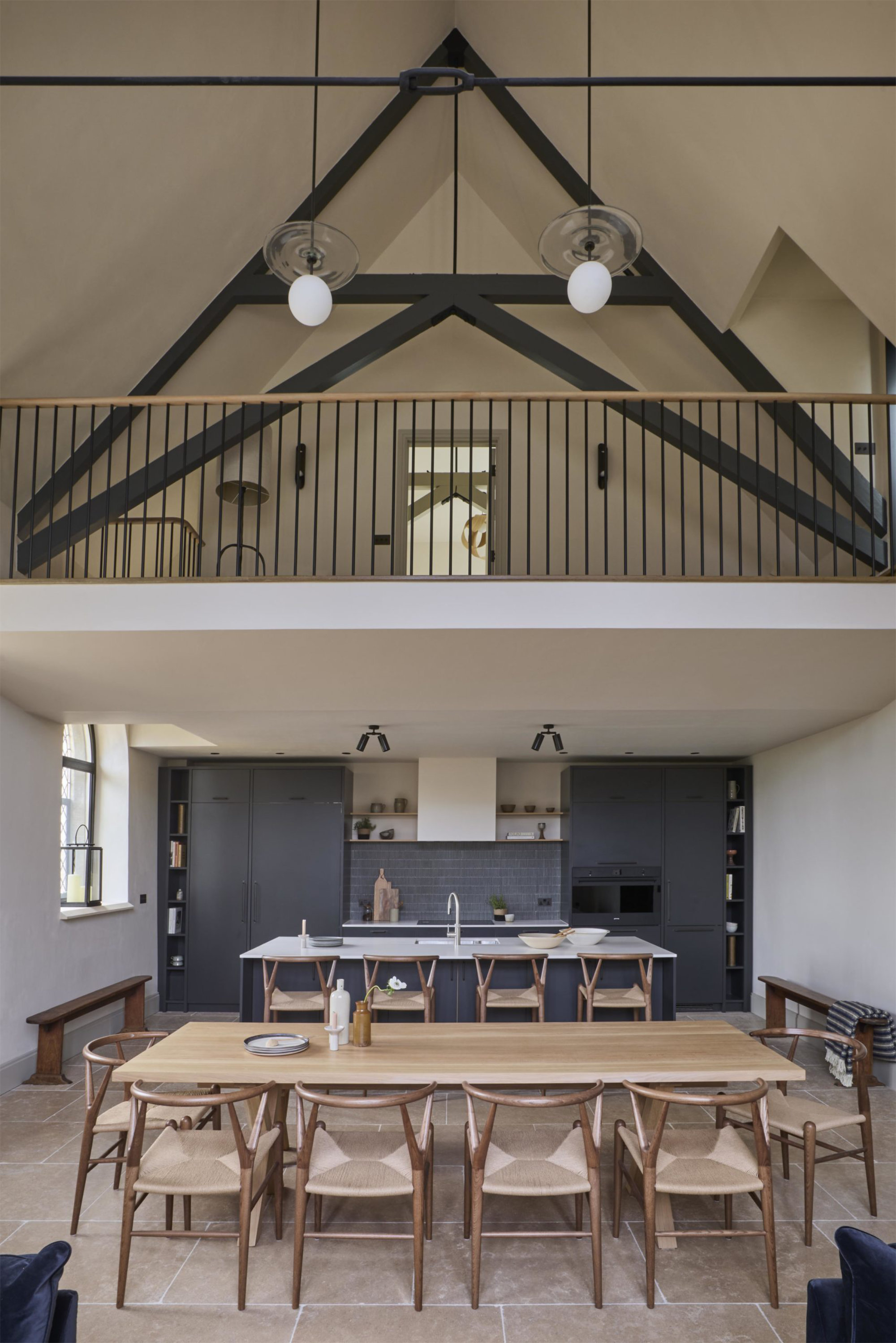 A double-height kitchen-diner in a chapel conversion with dramatic black steel roof trusses, a mezzanine gallery above, navy blue fitted kitchen, large oak dining table.