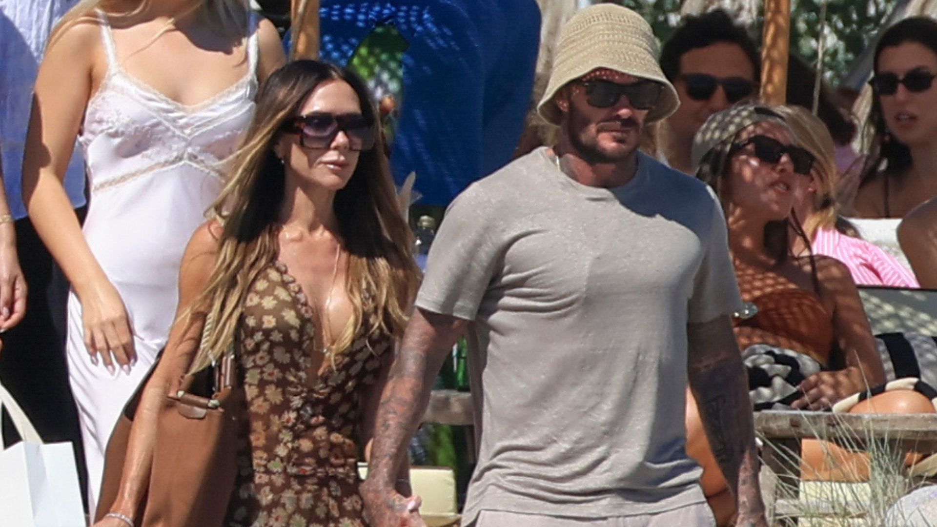Victoria and David Beckham walk along a beach holding hands. Victoria wears a brown sheer dress with Hermes Oran sandals whilst David wears a grey t-shirt with grey linen shorts.
