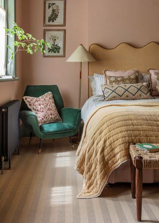 A neutral/ nude carpet with a double bed on top of it and a green accent chair with a pillow on top. There is also a window with a small vase and plant inside.