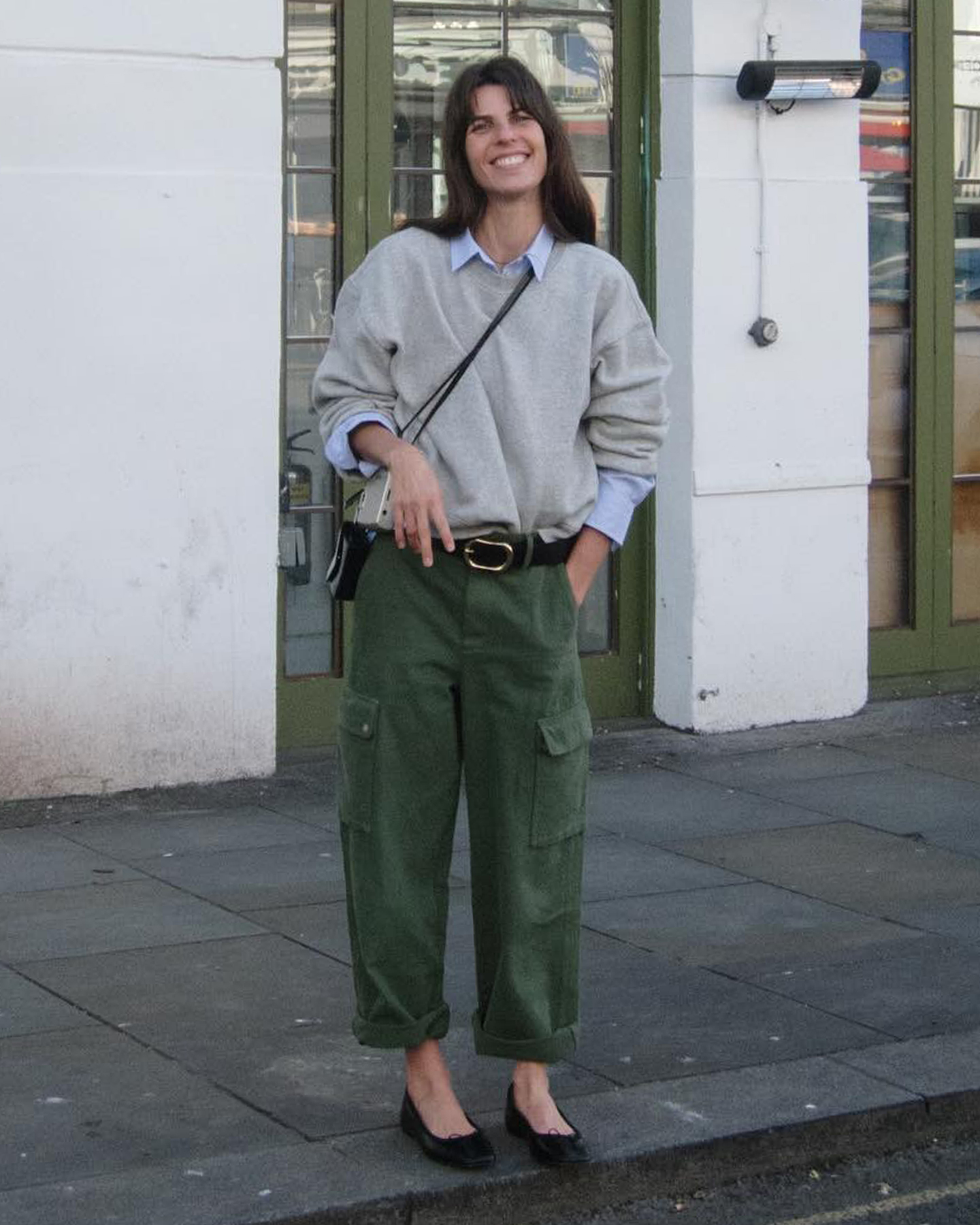 French style influencer Laura Vidrequin Roso poses in a gray sweatshirt layered over a blue button-down shirt, a black belt, crossbody bag, cuffed green cargo pants, and black flats