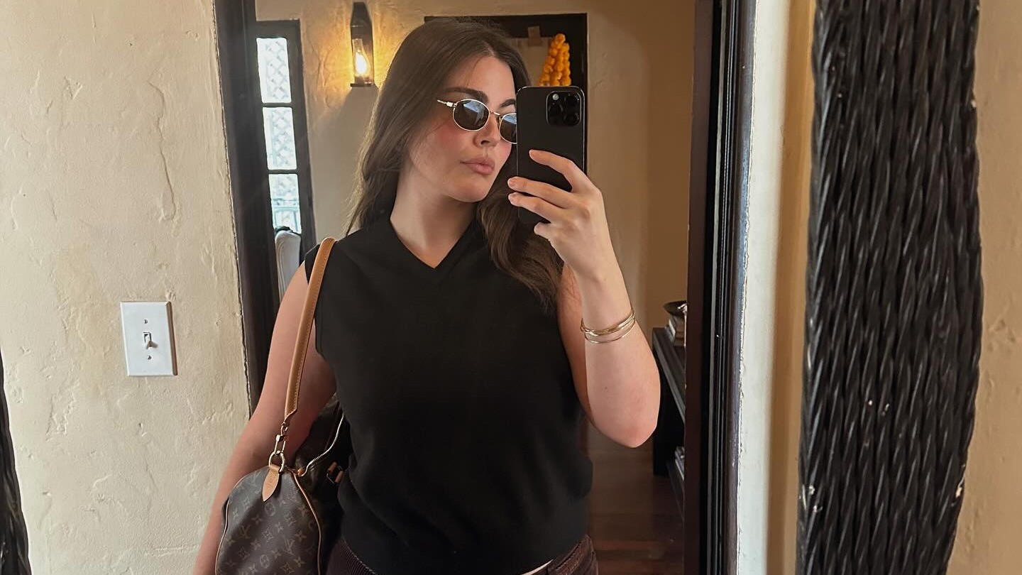 Woman wears v-neck black sweater vest with brown corduroys, a brown belt and round sunglasses.