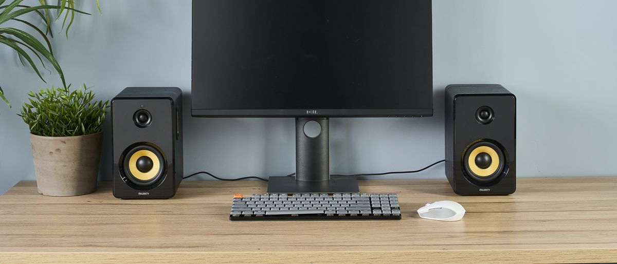 a pair of black wood speakers Majority D40X with a yellow kevlar cone photographed on a wood desk with a computer and blue wall and a plant in shot