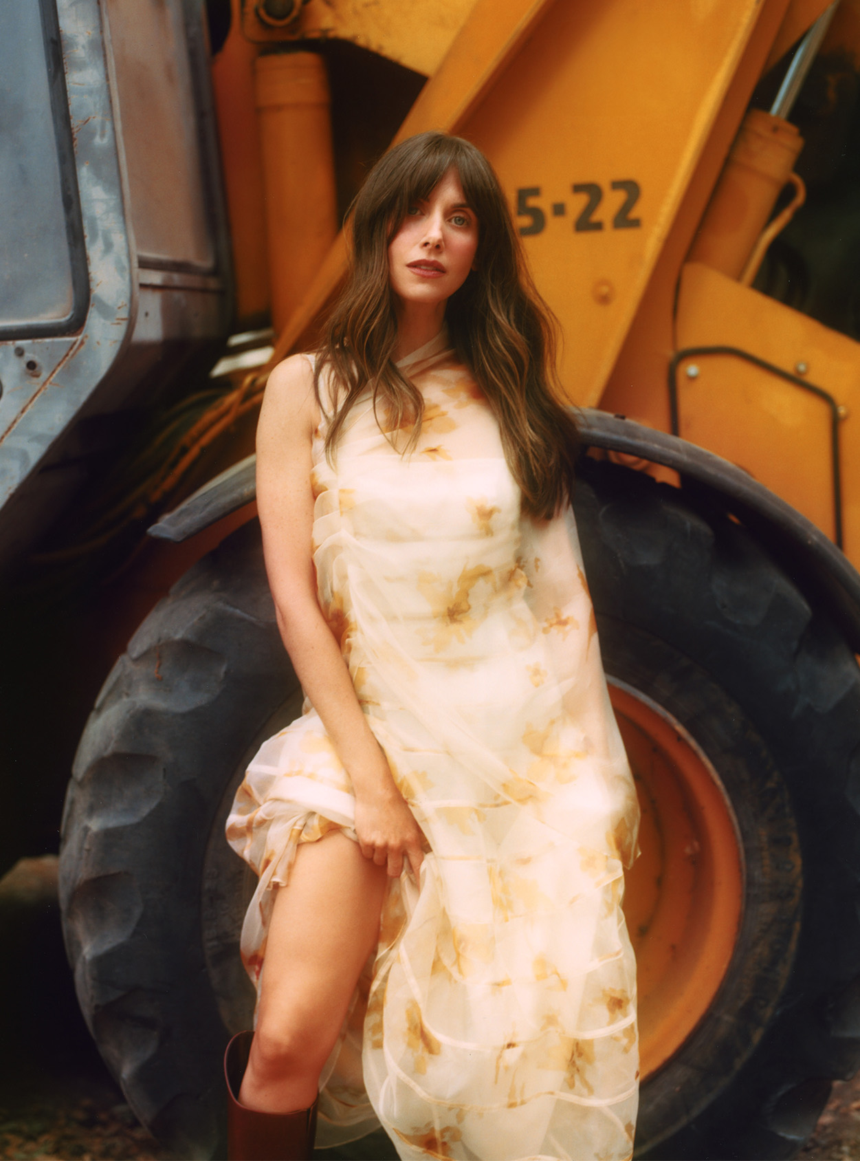Alison Brie stands in front of a bulldozer wearing a floral dress