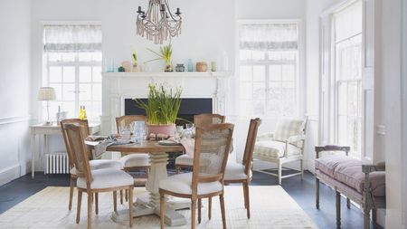 A bright spring dining room with a circular wooden dining table and rattan chairs in the center, and large bright windows all around with sheer curtains. In the back centre of the room is a large white mantelpiece with flowers and vases, and the table in the centre is set with daffodils, plates, and glasses. Beneath the table is a large cream rug, and to the right is a pale purple upholstered bench.