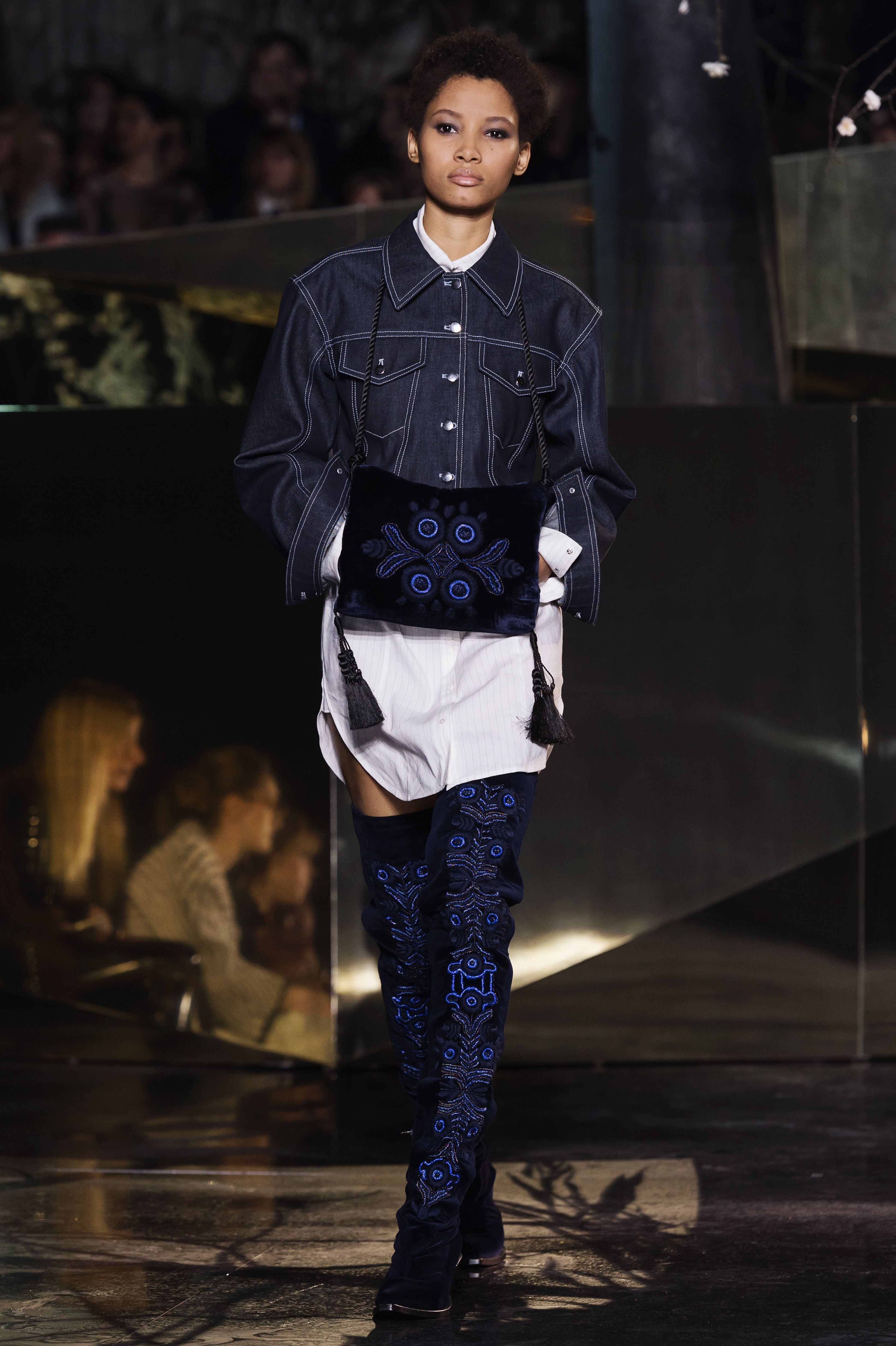 A model walks the runway wearing a denim jacket, white skirt, and thigh high boots.