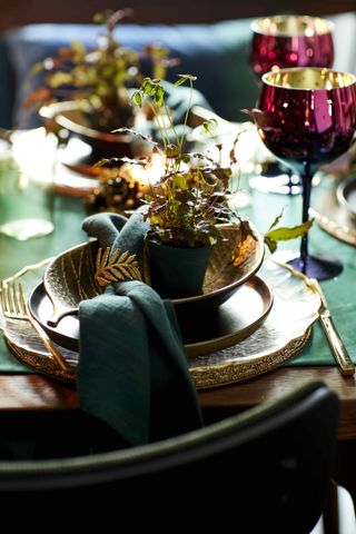 Christmas table setting with gold tableware, green napkin and runner, pink glasses, mini plant as a table gift