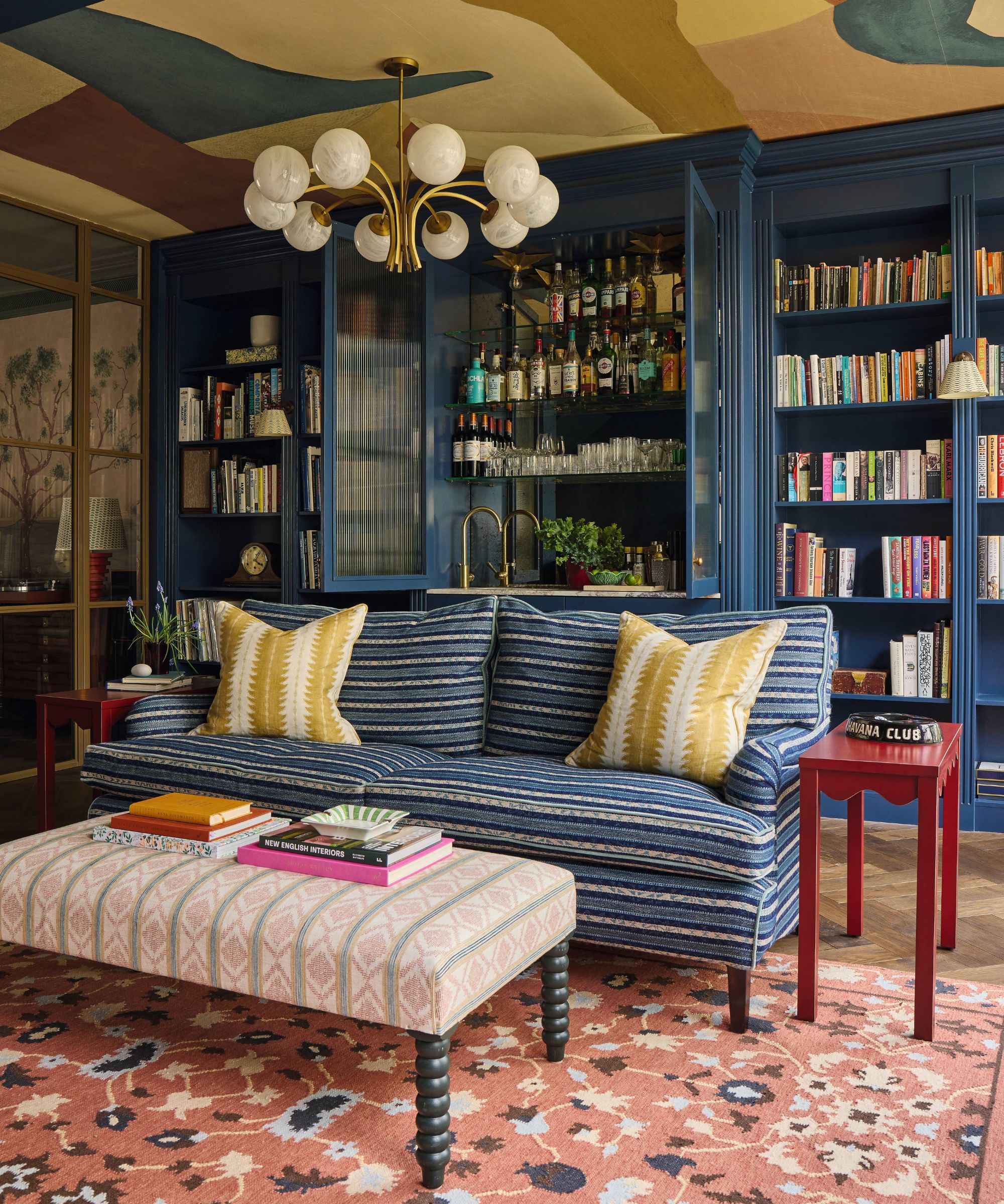 a snug TV room with custom blue cabinetry hiding a home bar with a wallpapered ceiling, striped blue couch with yellow pillows and a rust red vintage rug and striped ottoman