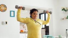 woman in a white kitchen background holding up a light dumbbell in each hand to either side of her at head height facing the camera diagonally wearing a yellow long sleeved zip neck top 