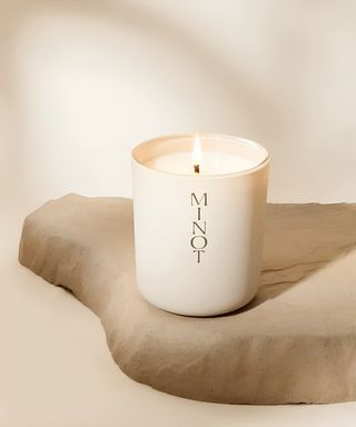 A cream glass jar with a white candle burning inside, on a stone slab against a matching cream background.